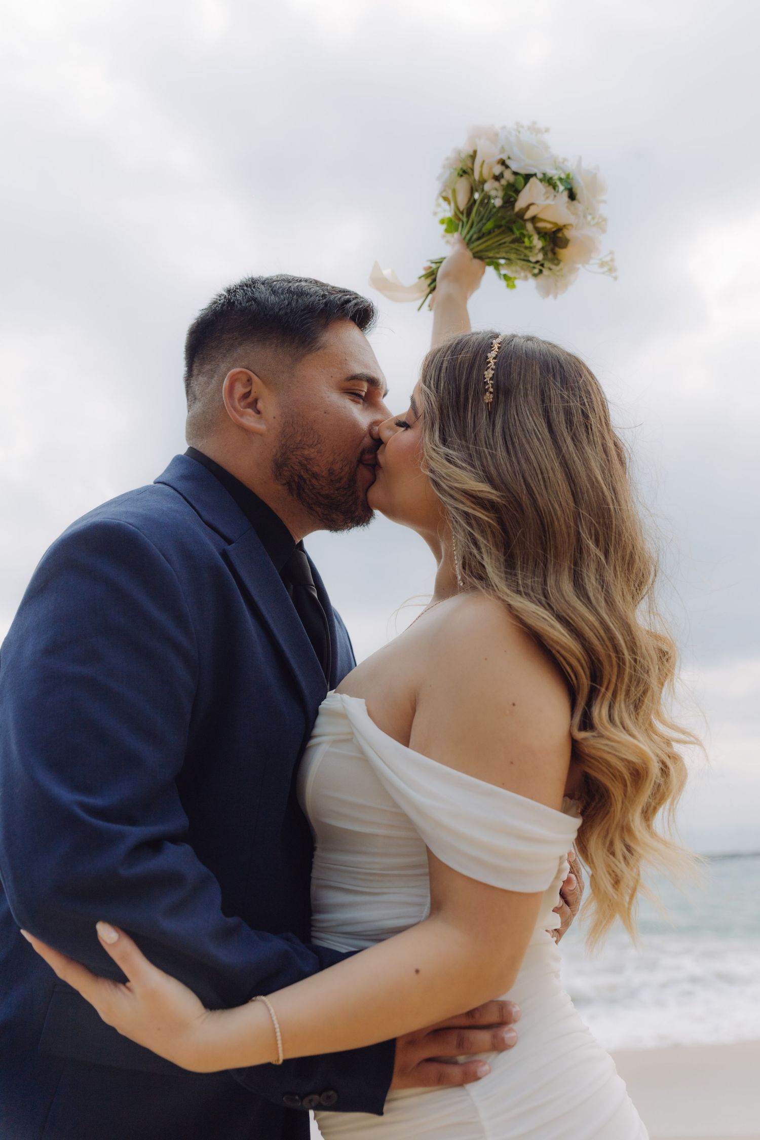 Bride and groom sharing a romantic kiss on the beach, bride raising white floral bouquet overhead.