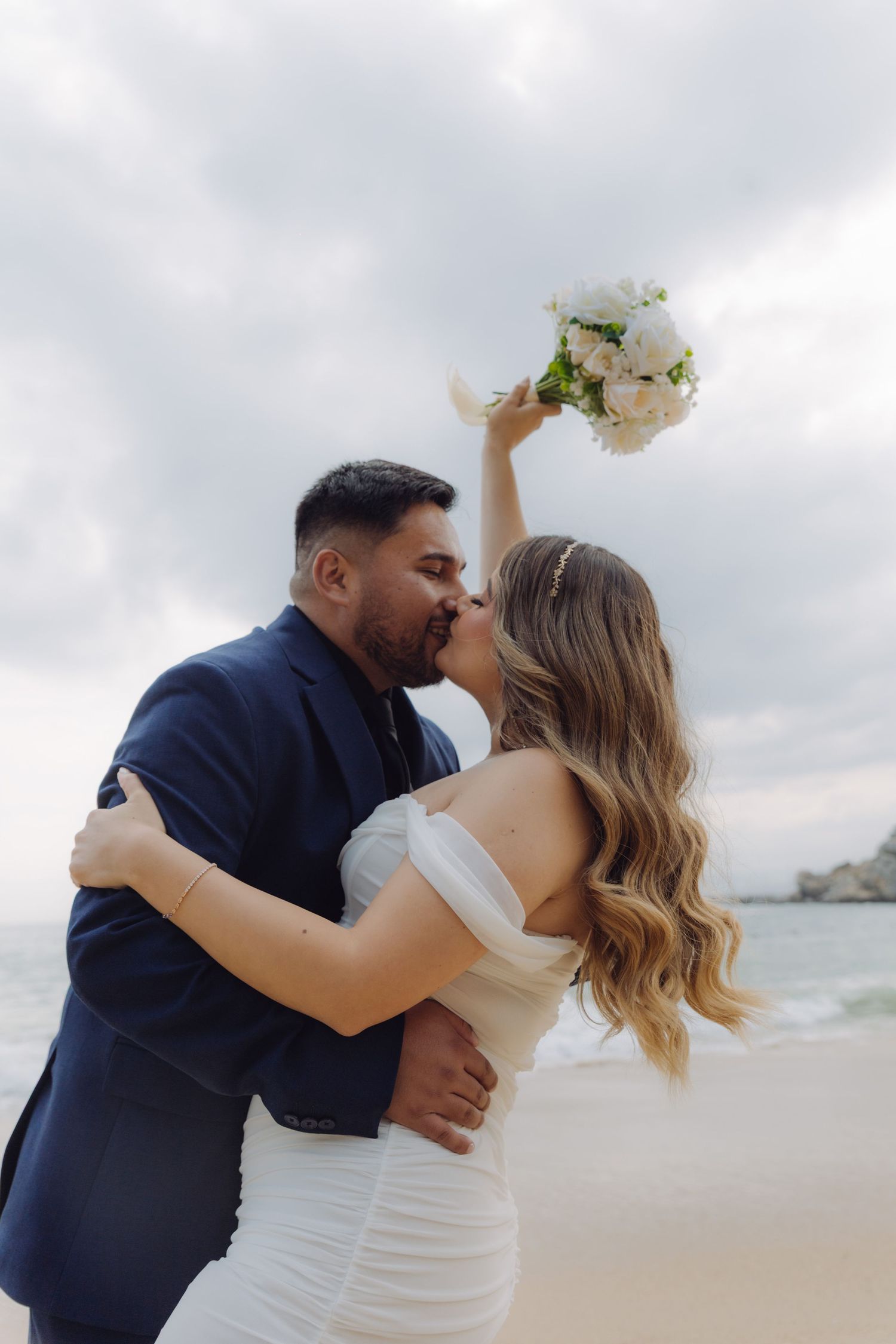 Bride and groom share a kiss on the beach, bride holding white floral bouquet overhead in wedding attire.
