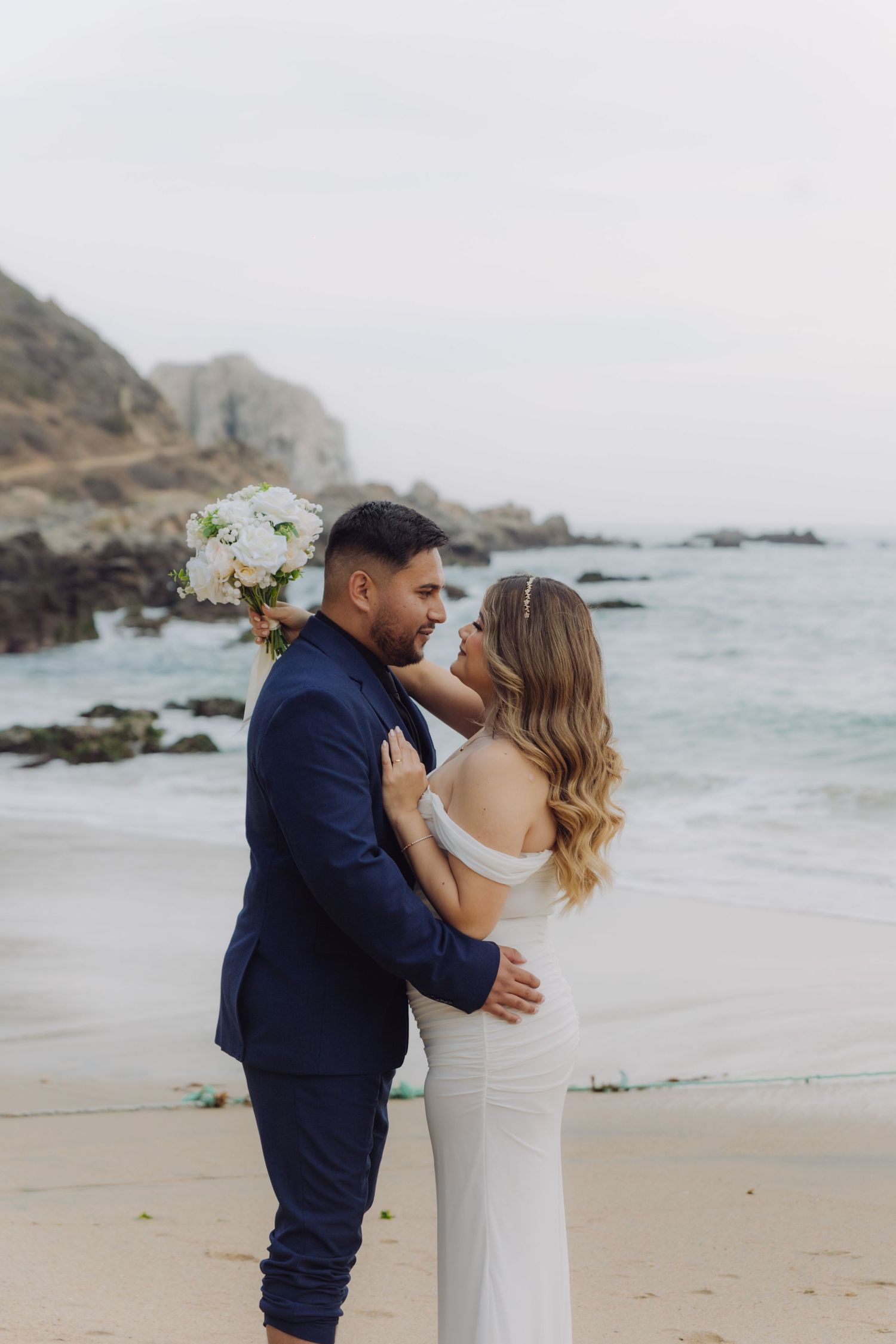 Bride and groom embrace on sandy beach, groom in navy suit, bride in white off-shoulder gown with bouquet.