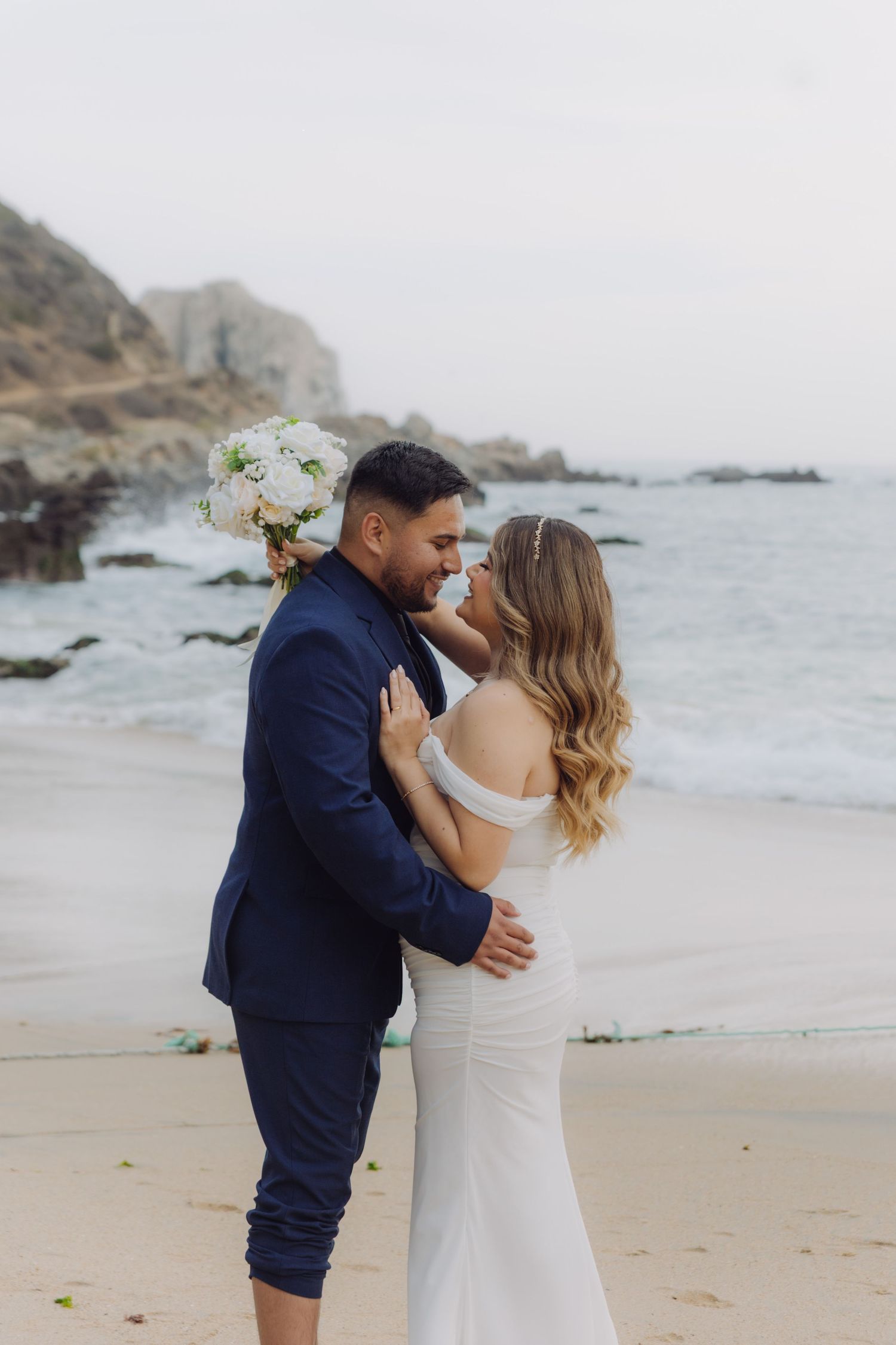 Newlyweds share a romantic kiss on a rocky beach, bride in fitted white gown holding white flower bouquet.