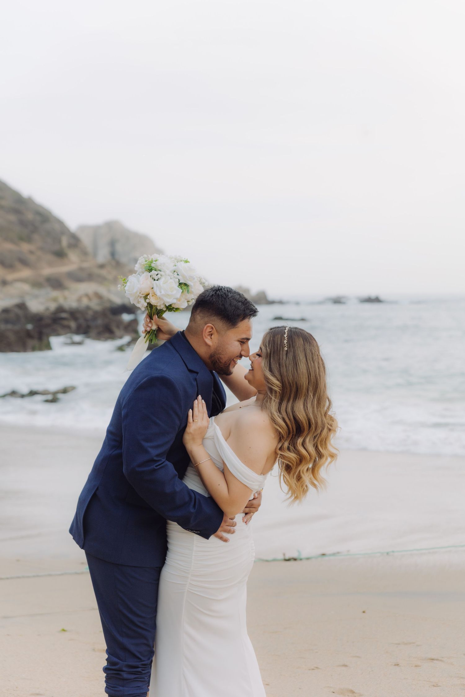Bride and groom lean in for a kiss on a scenic beach, surrounded by rocky cliffs and ocean waves.