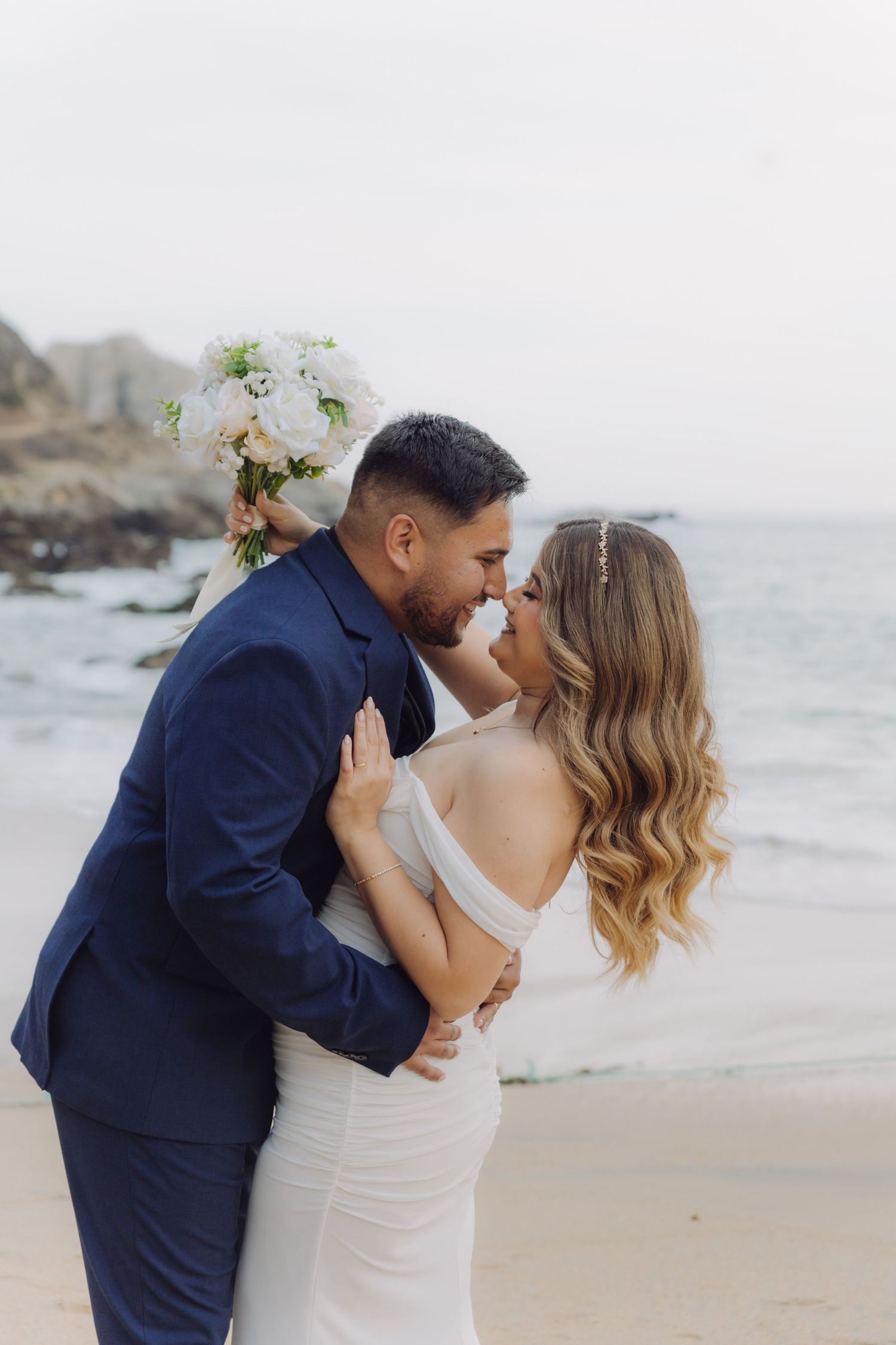 Couple shares an intimate kiss on the beach, bride in elegant white gown, groom in navy blue suit.