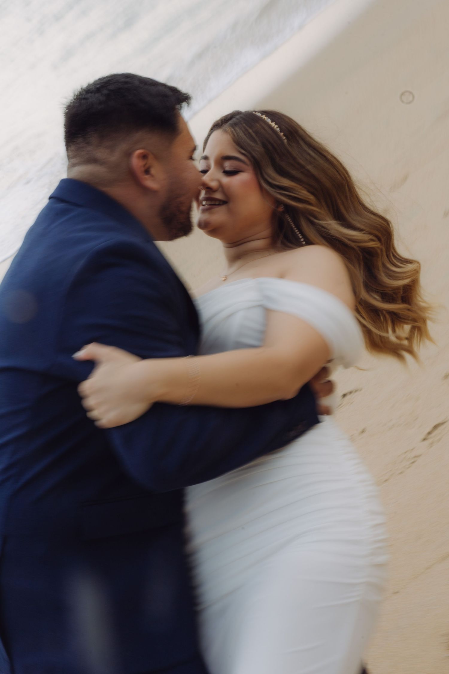 Bride and groom share a joyful embrace, bride in white off-shoulder fitted gown with bridal hair accessory.