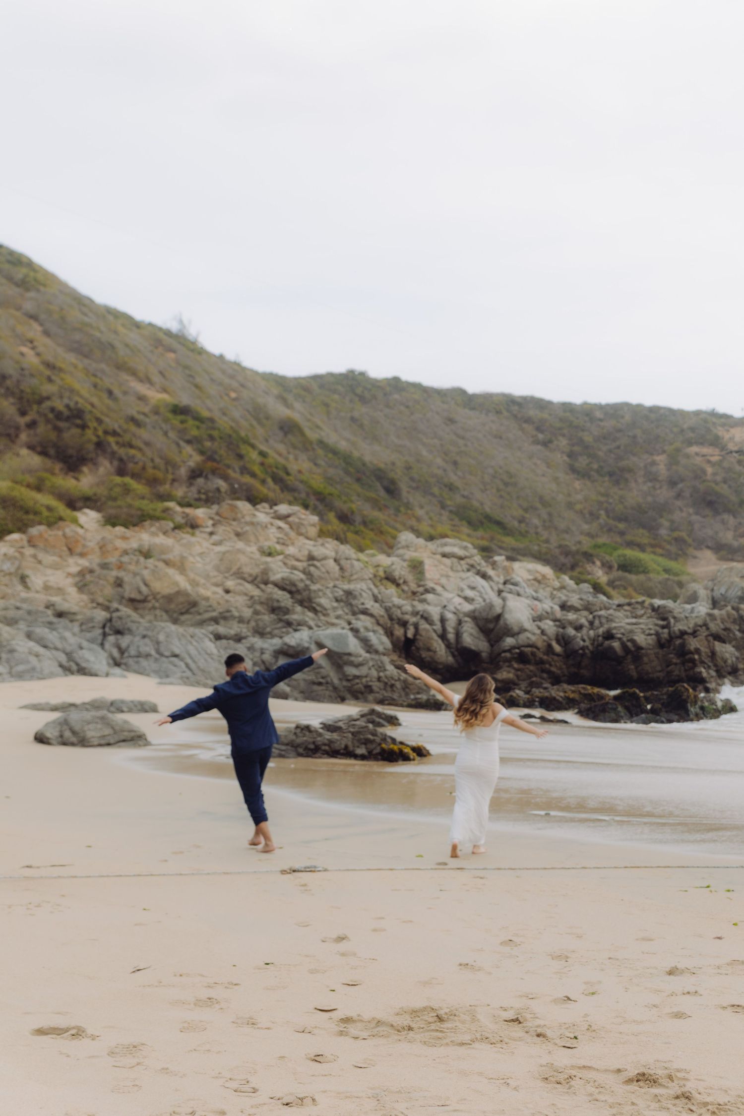 Bride and groom dance playfully on a sandy beach with rocky cliffs and greenery in the background.