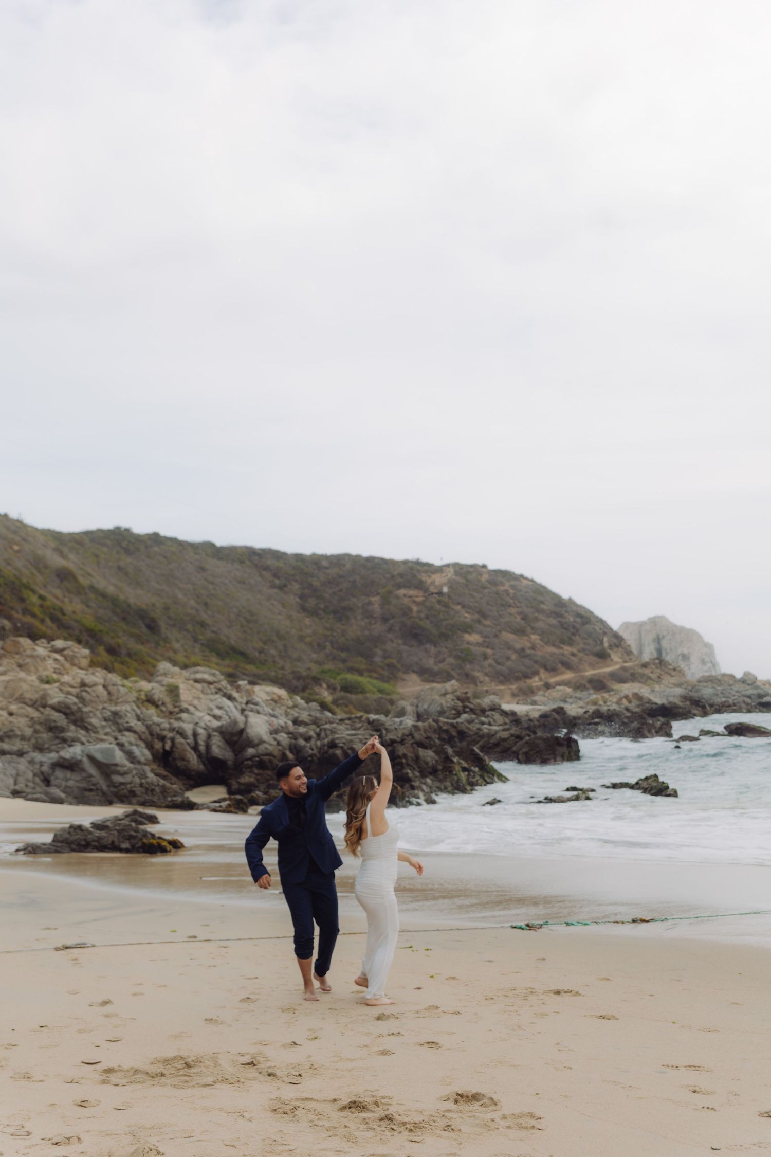 Newlyweds dance joyfully on a scenic beach, groom in navy suit and bride in flowing white wedding gown.