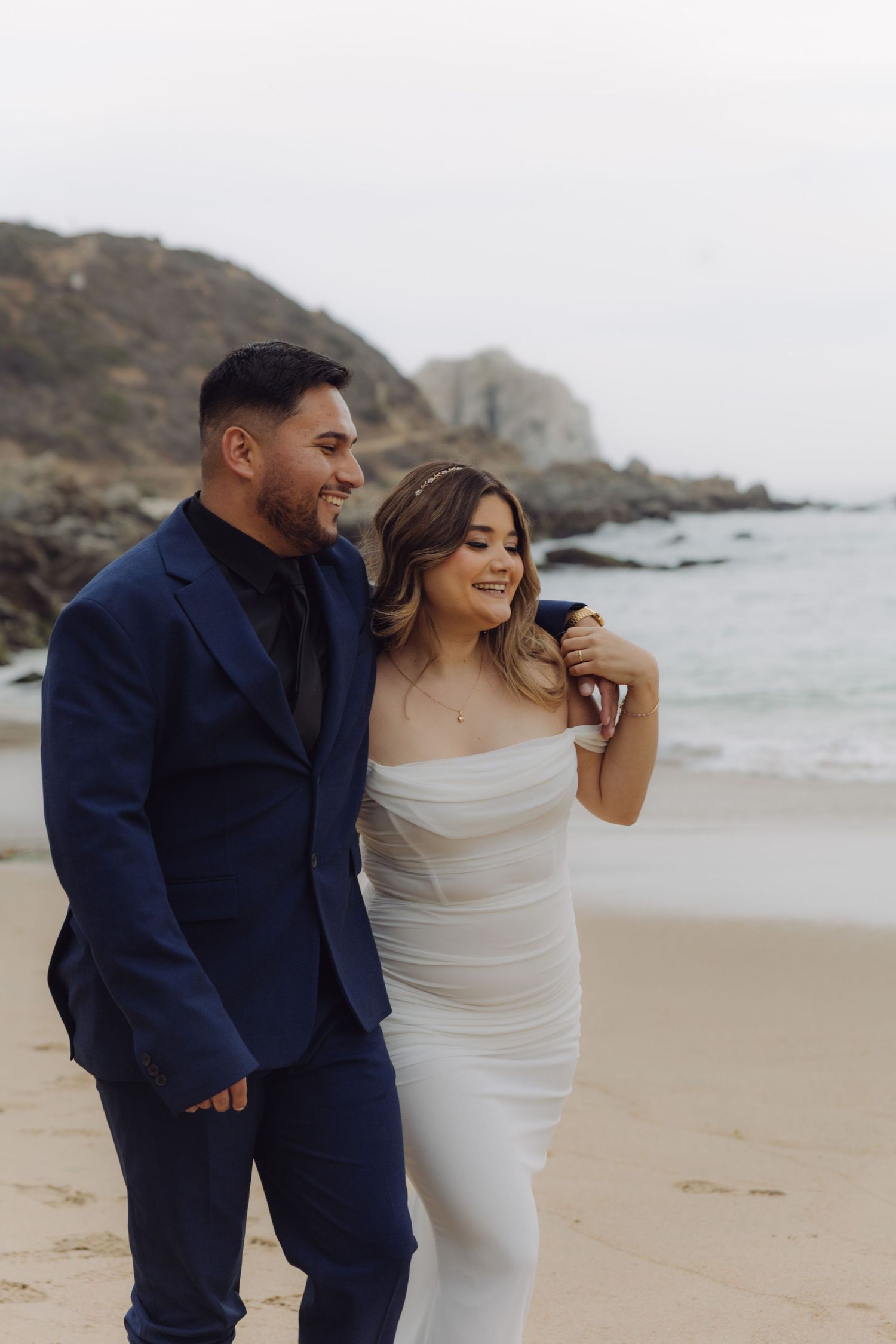 Smiling bride and groom walk together on a beach, bride in white strapless gown, groom in navy suit.