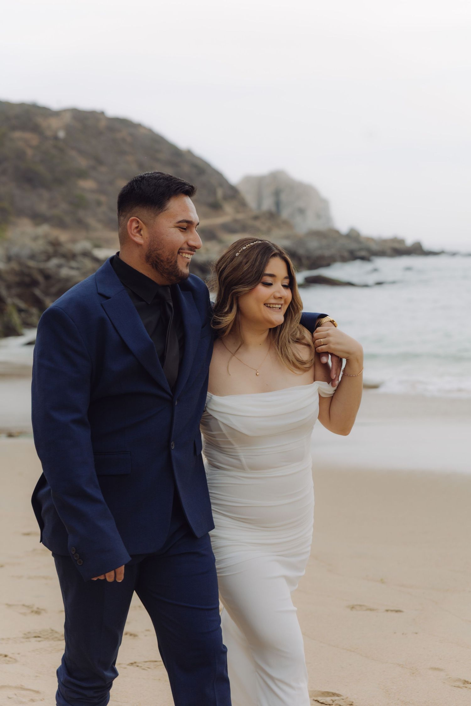 Happy couple strolls along a rocky beach shoreline, bride in white off-shoulder gown, groom in navy suit.