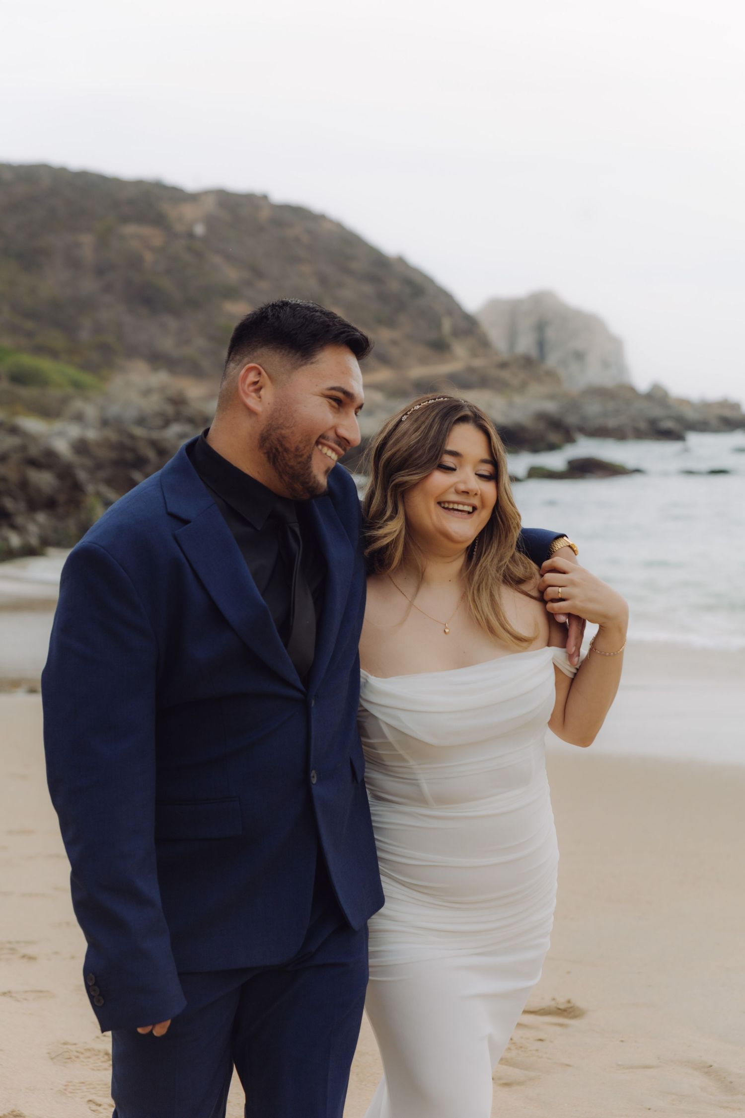 Smiling couple in wedding attire walking on a sandy beach, groom in navy suit, bride in white strapless gown.