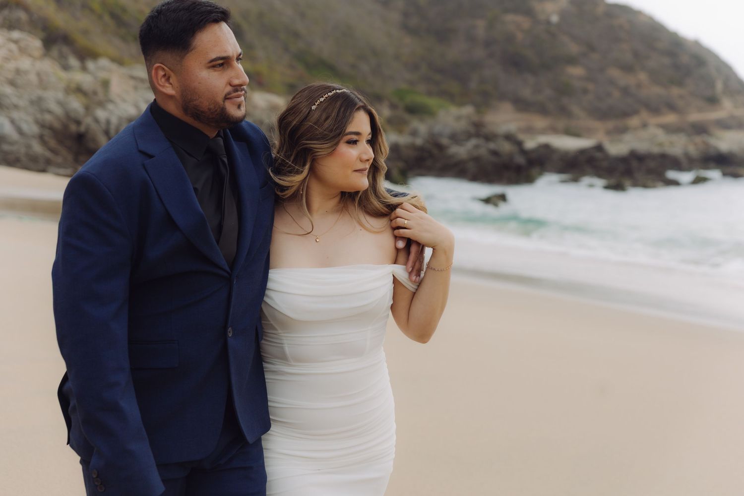 Wedding couple posing on a beach, groom in navy blue suit embracing bride in white strapless dress with rocky cliffs.