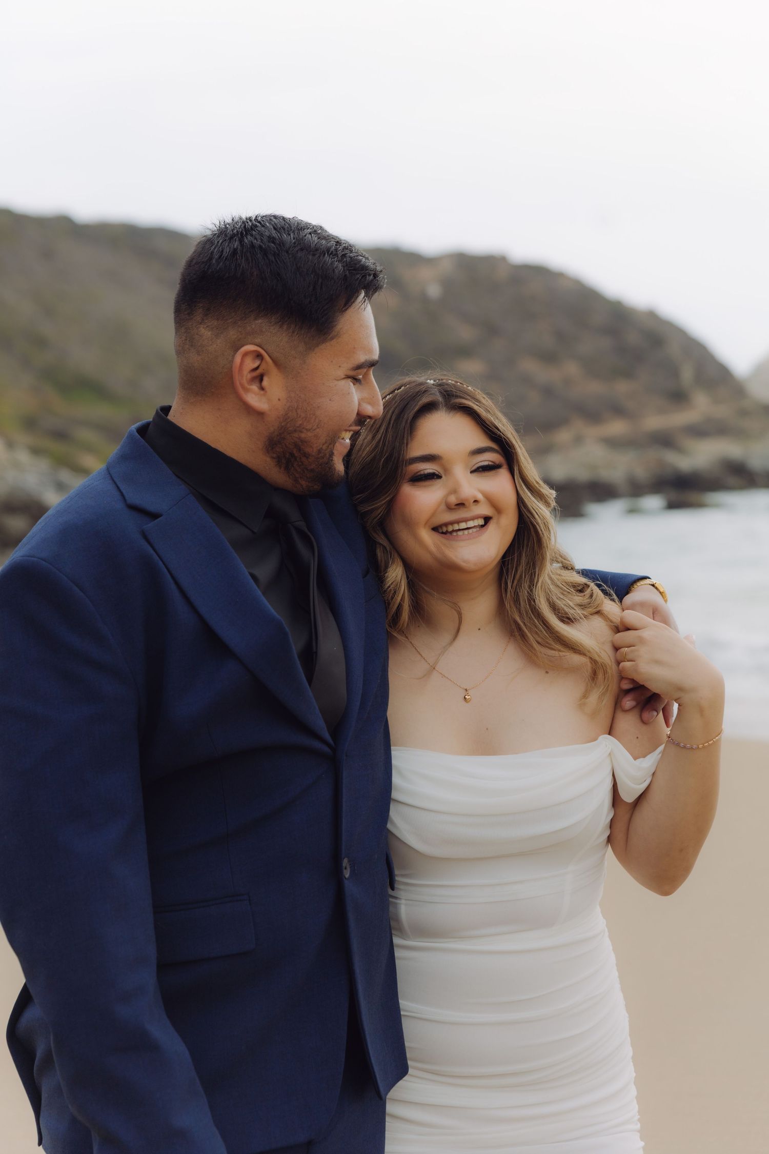 Happy wedding couple sharing a tender moment on a sandy beach, groom in navy suit and bride in white dress.