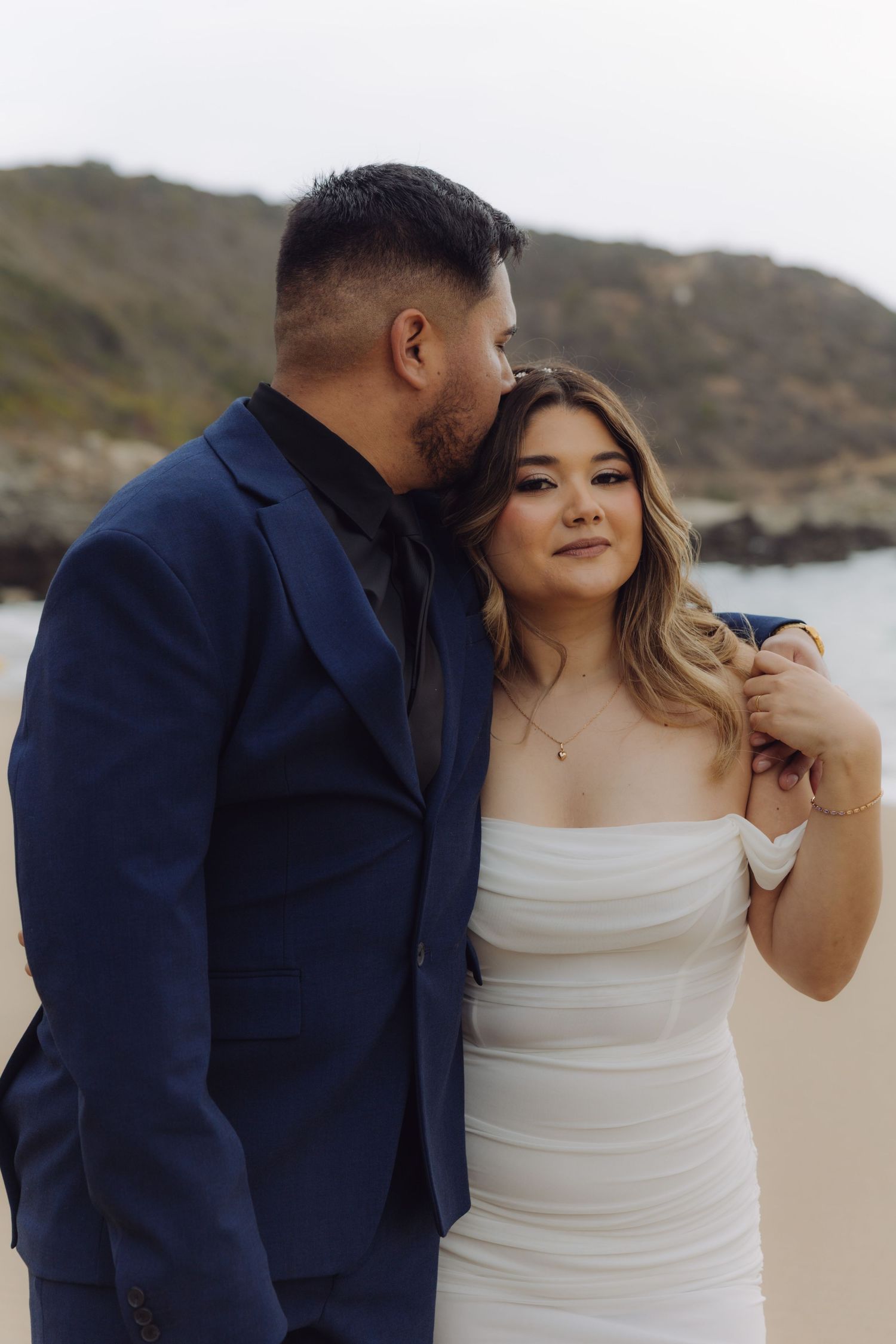 Groom in navy suit gently kissing brides temple as she smiles softly on a picturesque beach setting.