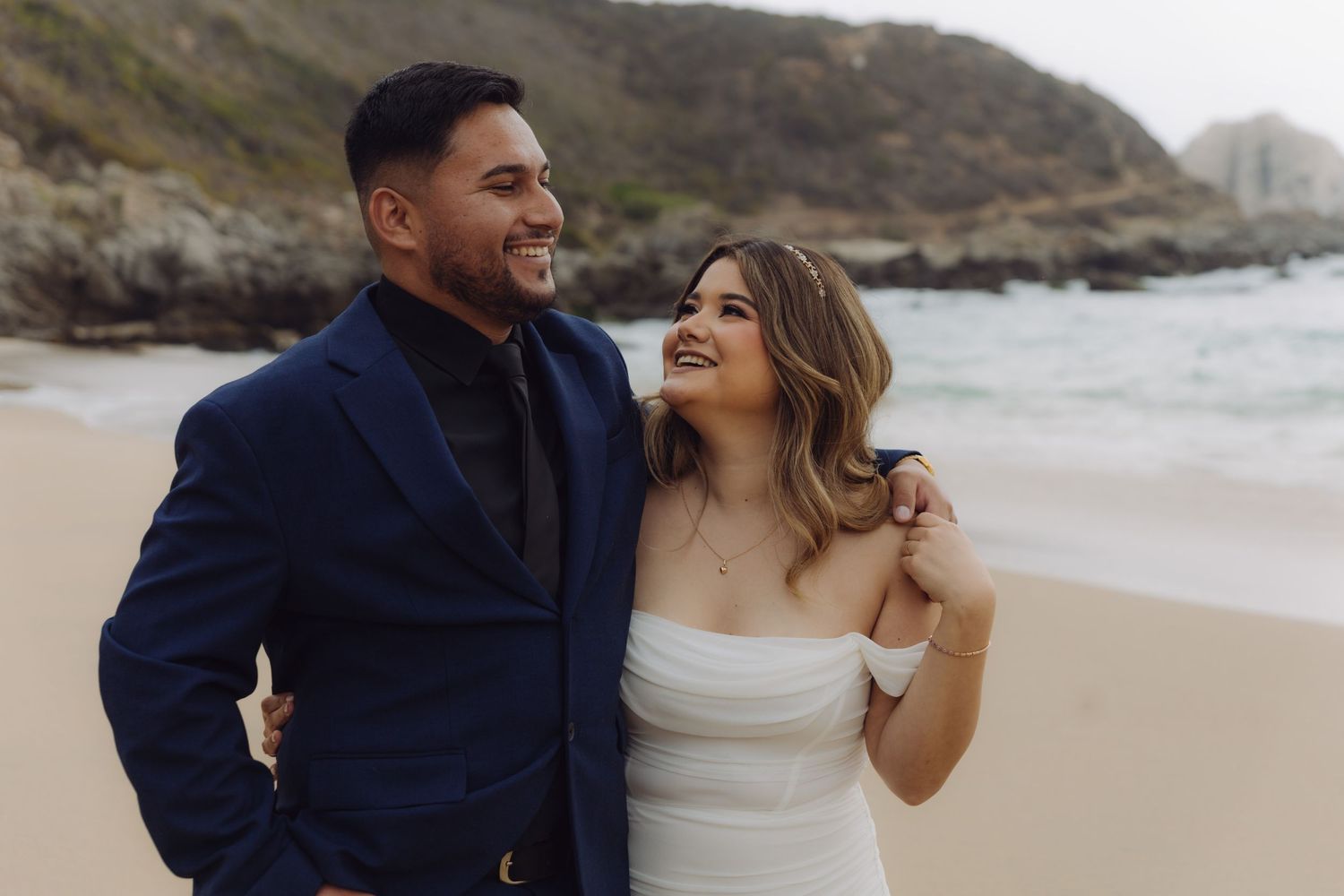 Elegant wedding couple smiling together on a sandy beach, groom in navy suit and bride in white strapless gown.