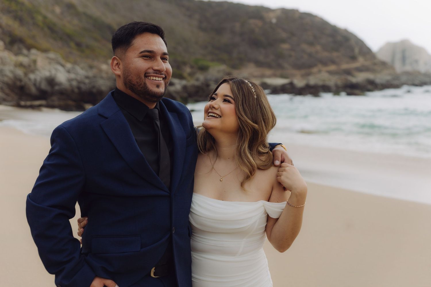Bride and groom posing on a beach, bride in white strapless dress looking up at groom in navy blue suit.