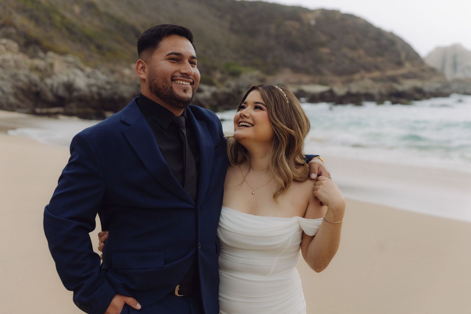 Smiling wedding couple standing on a sandy beach with rocky coastal cliffs in the background.