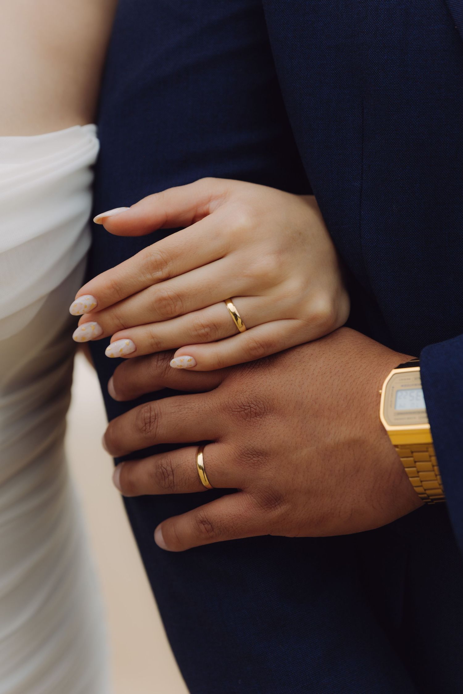 Close-up of newlyweds hands showing matching gold wedding bands, bride with nude manicure and groom wearing gold watch.