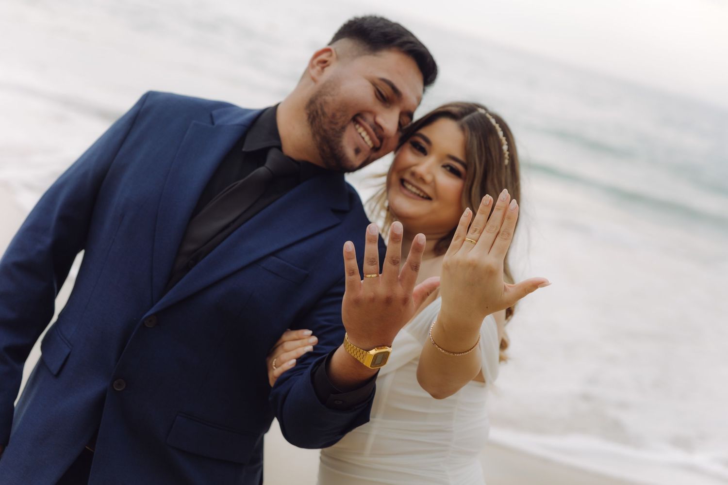 Smiling newlyweds showing off their wedding rings on a beach, groom in navy suit, bride in white.