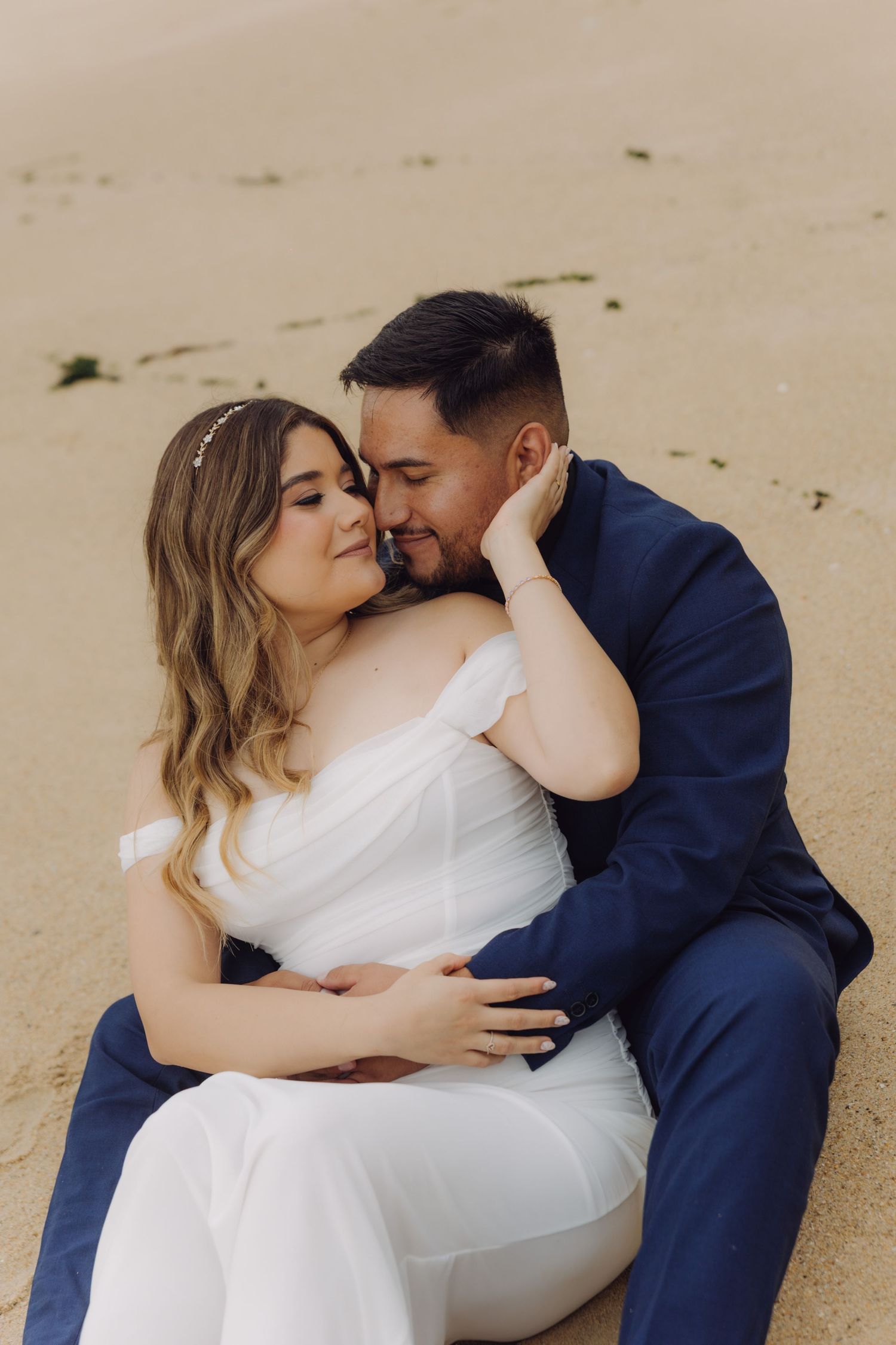 Newlyweds in wedding attire sitting on sandy beach, bride in white dress and groom in navy suit.