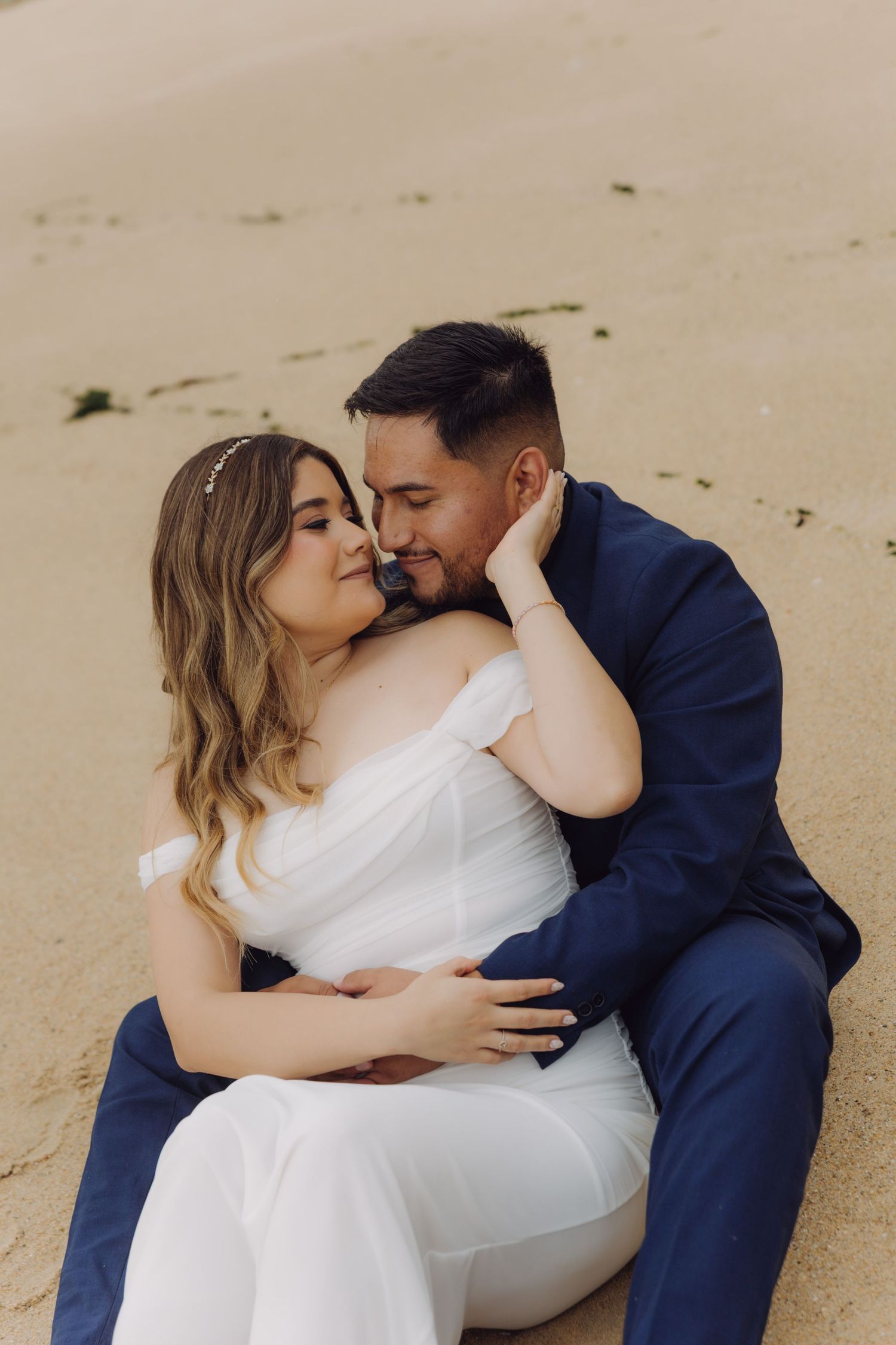 Bride and groom sitting closely on sandy beach, sharing a romantic embrace in wedding attire.