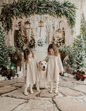 Two young girls in matching plaid dresses pose with a white dog amid festive Christmas trees and lanterns.
