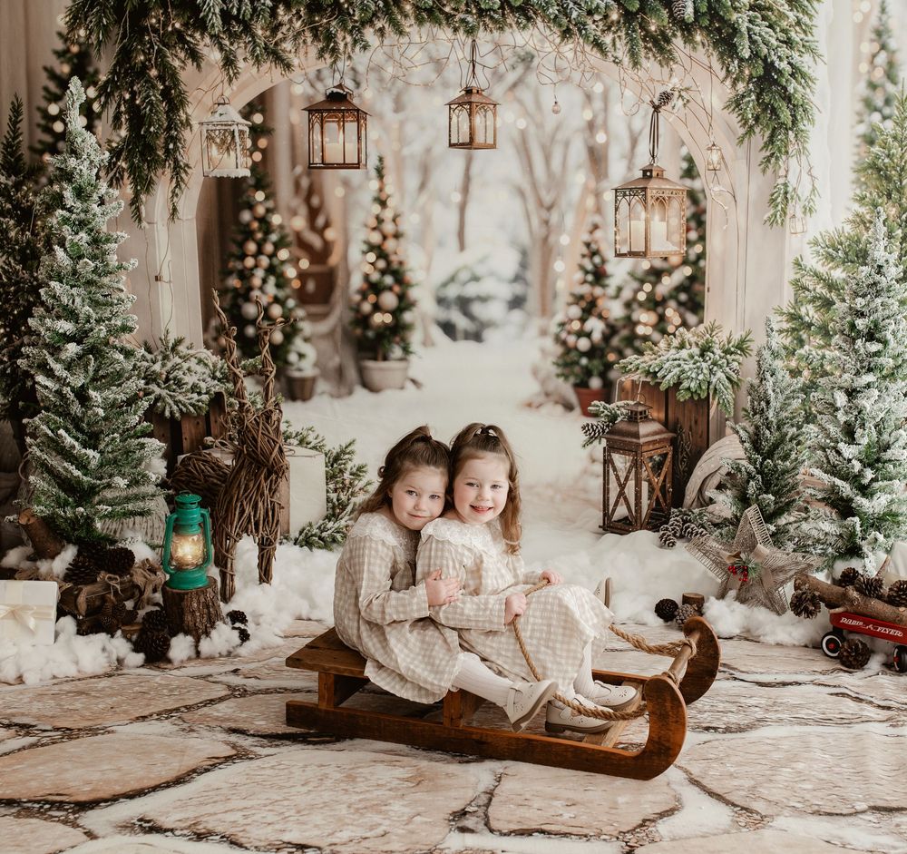 Two young girls in matching outfits sit together on a wooden sled in a festive winter Christmas photo backdrop.