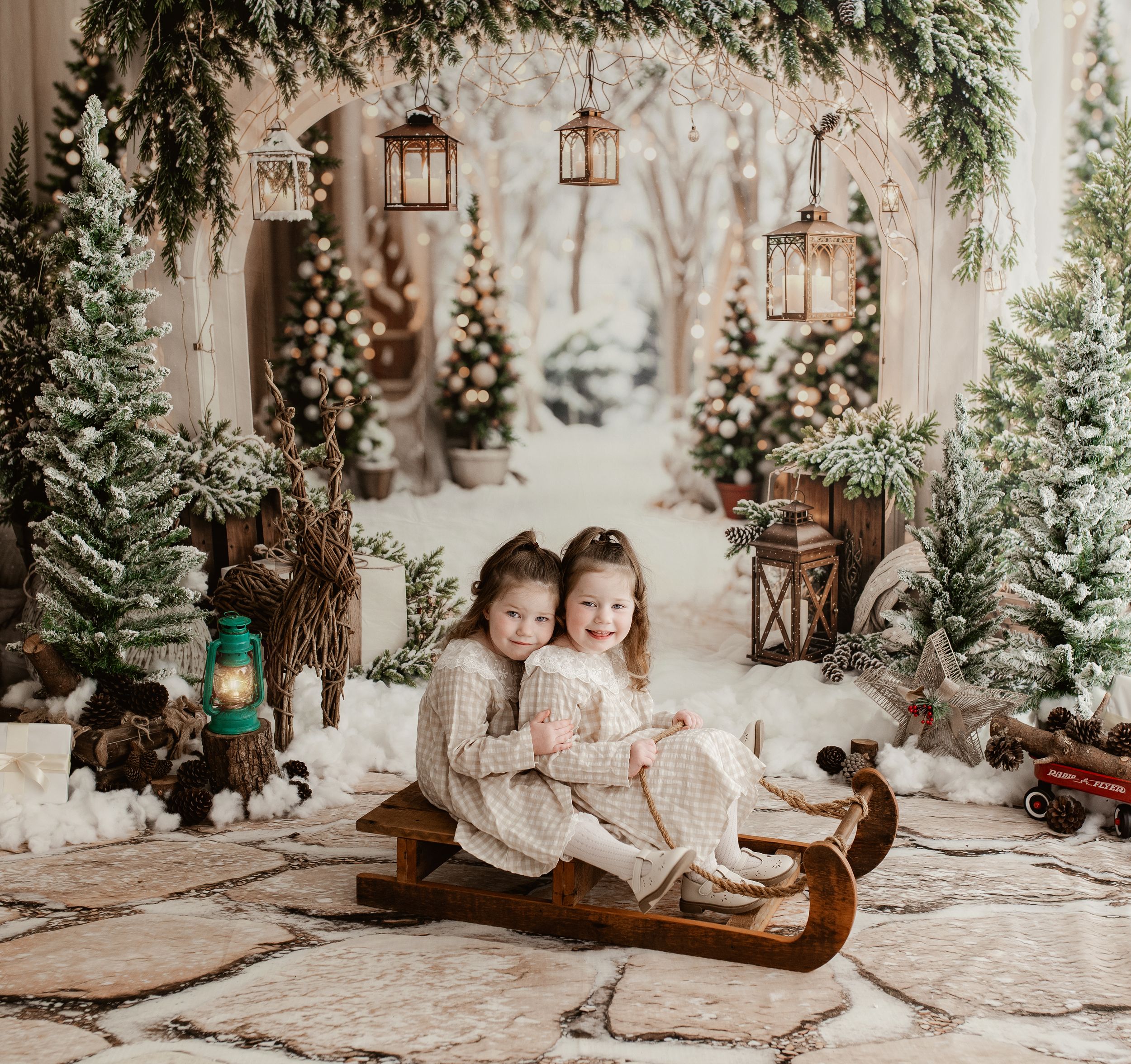 Two young girls in matching outfits sit together on a wooden sled in a festive winter Christmas photo backdrop.