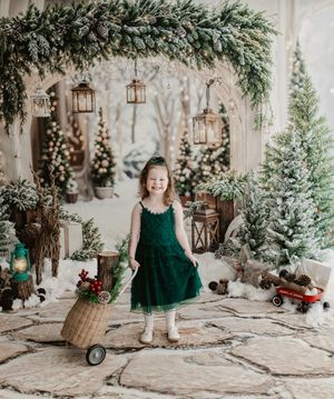 Young girl in green dress poses with wicker cart amid festive Christmas decor, snow-dusted trees, and lanterns.