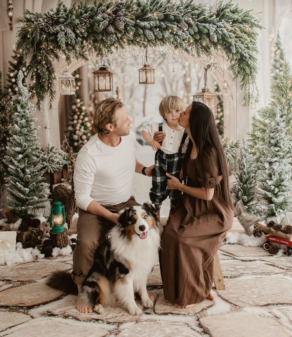 Family with young child and Australian Shepherd dog pose in festive Christmas setting with snow-dusted trees.
