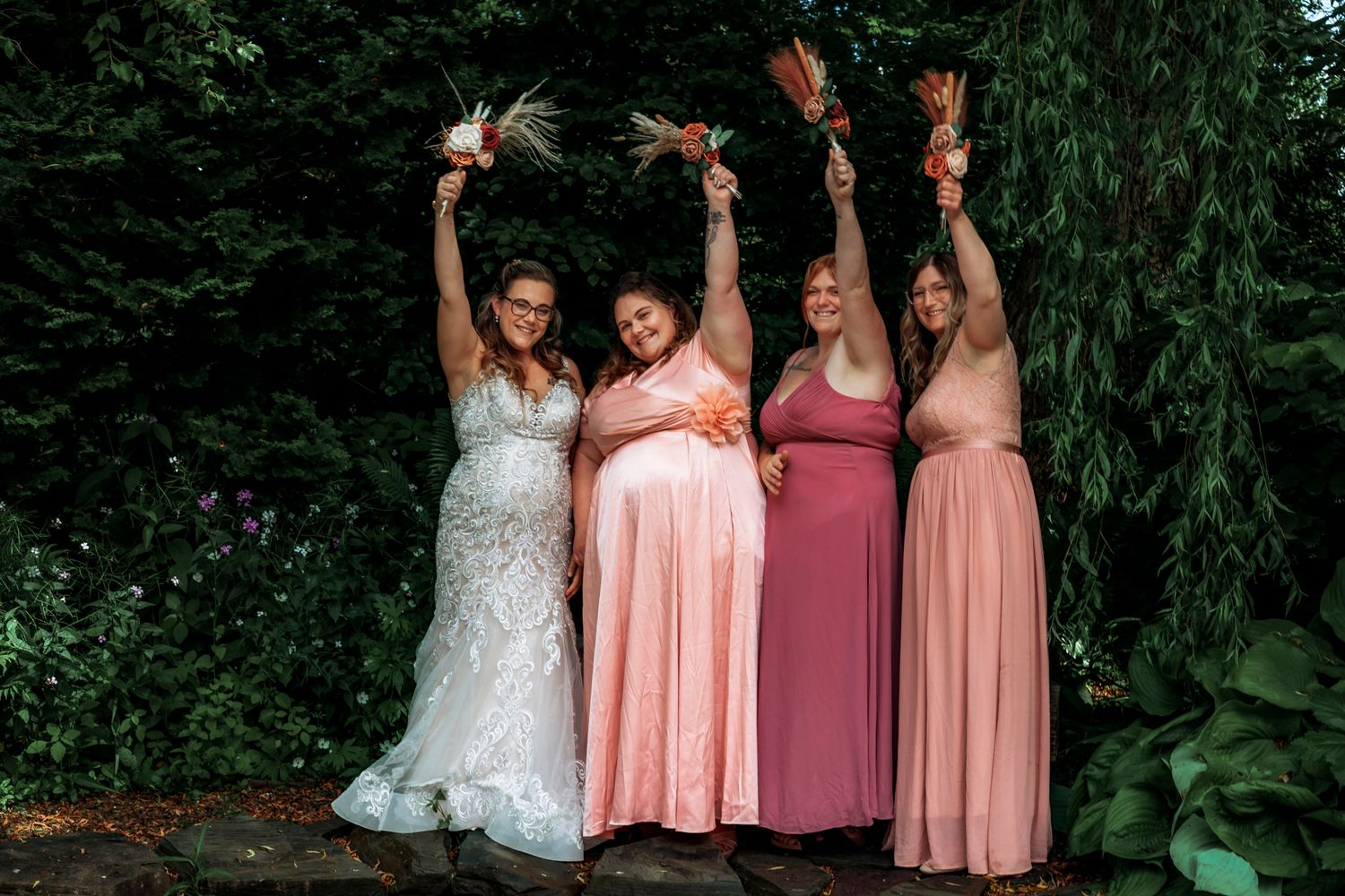 Bride in white lace gown with three bridesmaids in pink dresses raising bouquets outdoors at wedding.