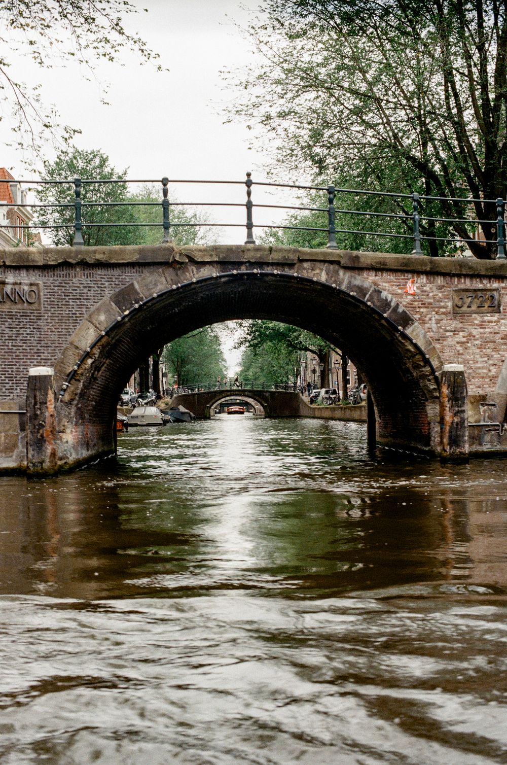 Historic brick arch bridge over Amsterdam canal with reflective water and lush green trees in background.