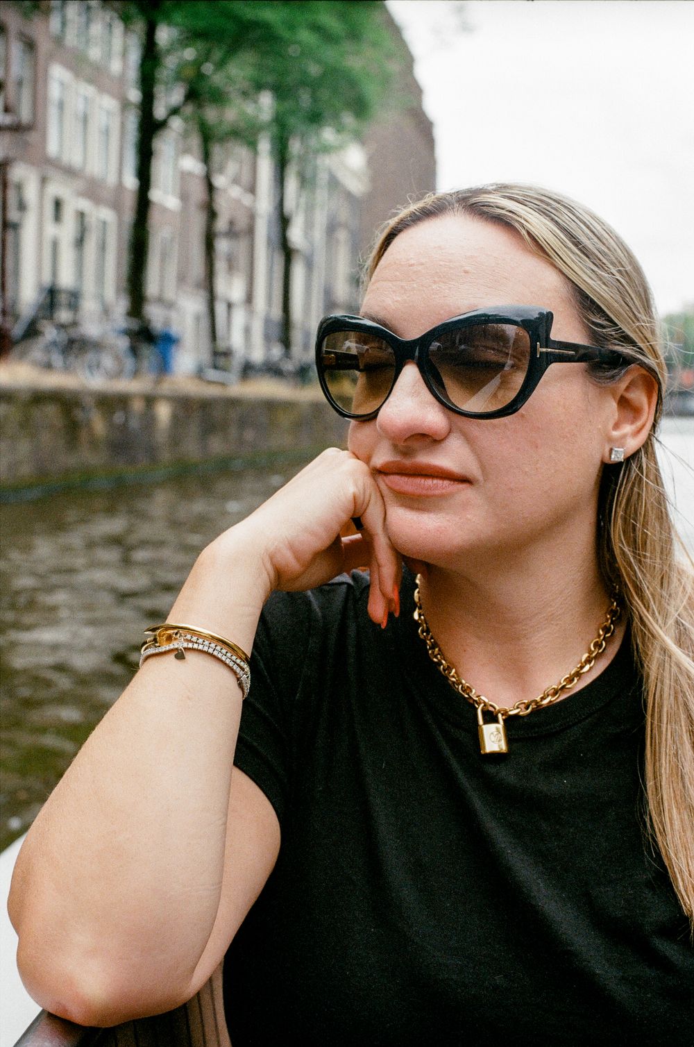 Woman in black cat-eye sunglasses and gold chain necklace sitting by Amsterdam canal on overcast day.