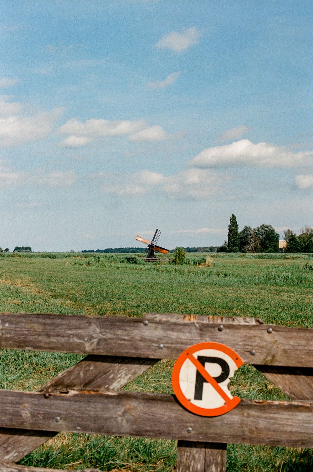 No parking sign on wooden fence in Dutch countryside with traditional windmill and green fields under blue sky.