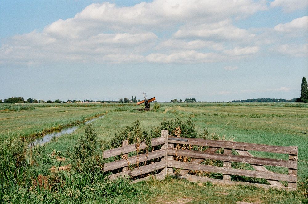 A wooden fence overlooks a vast green Dutch polder landscape with a windmill and canal under a cloudy sky.