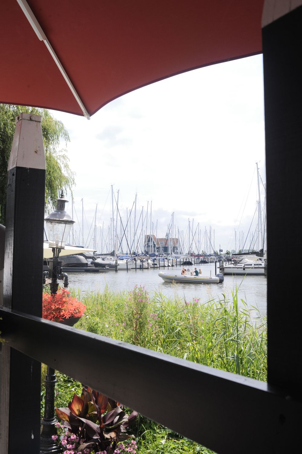 View from a shaded terrace overlooking a marina with sailboats, lush greenery, and a motorboat on calm water.