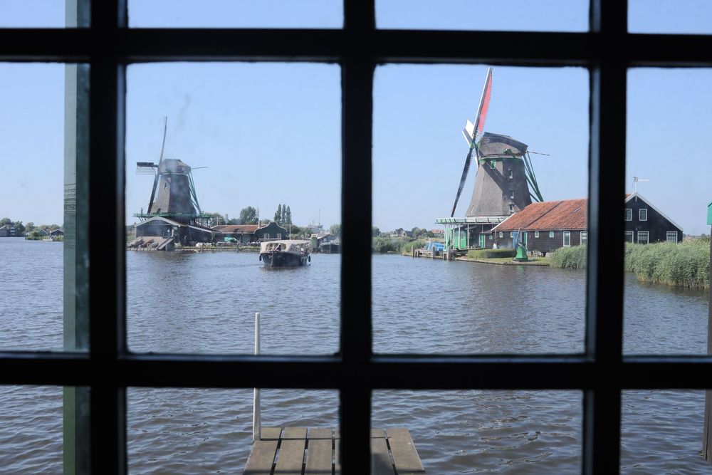 View through a window of Dutch windmills along a tranquil river in Zaanse Schans, Netherlands, with a boat passing by.