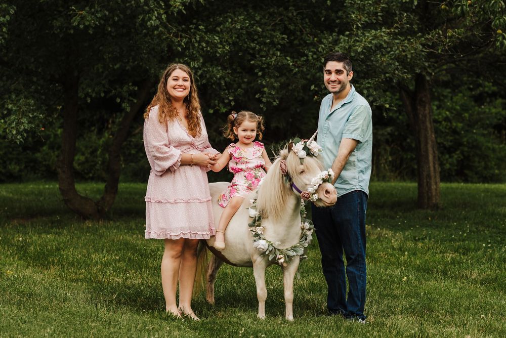 Family portrait with young girl sitting on a decorated white pony, flanked by smiling parents in a lush green park.