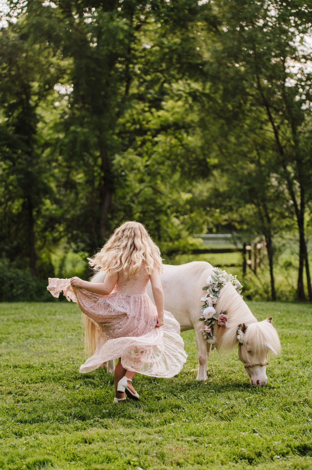 A young girl in a pink dress runs beside a white pony adorned with a floral wreath on a lush green lawn.