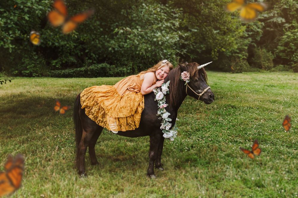 Girl in golden dress hugging a dark pony with floral garland, surrounded by monarch butterflies in a lush green meadow.