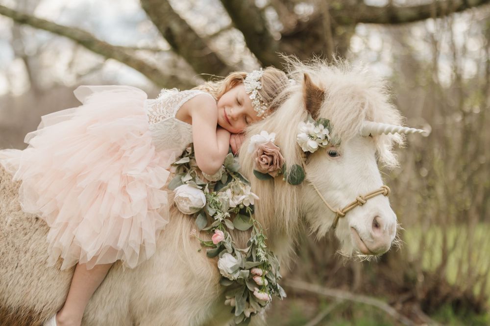 A little girl in a pink tutu dress rests on a flower-adorned white unicorn pony outdoors.