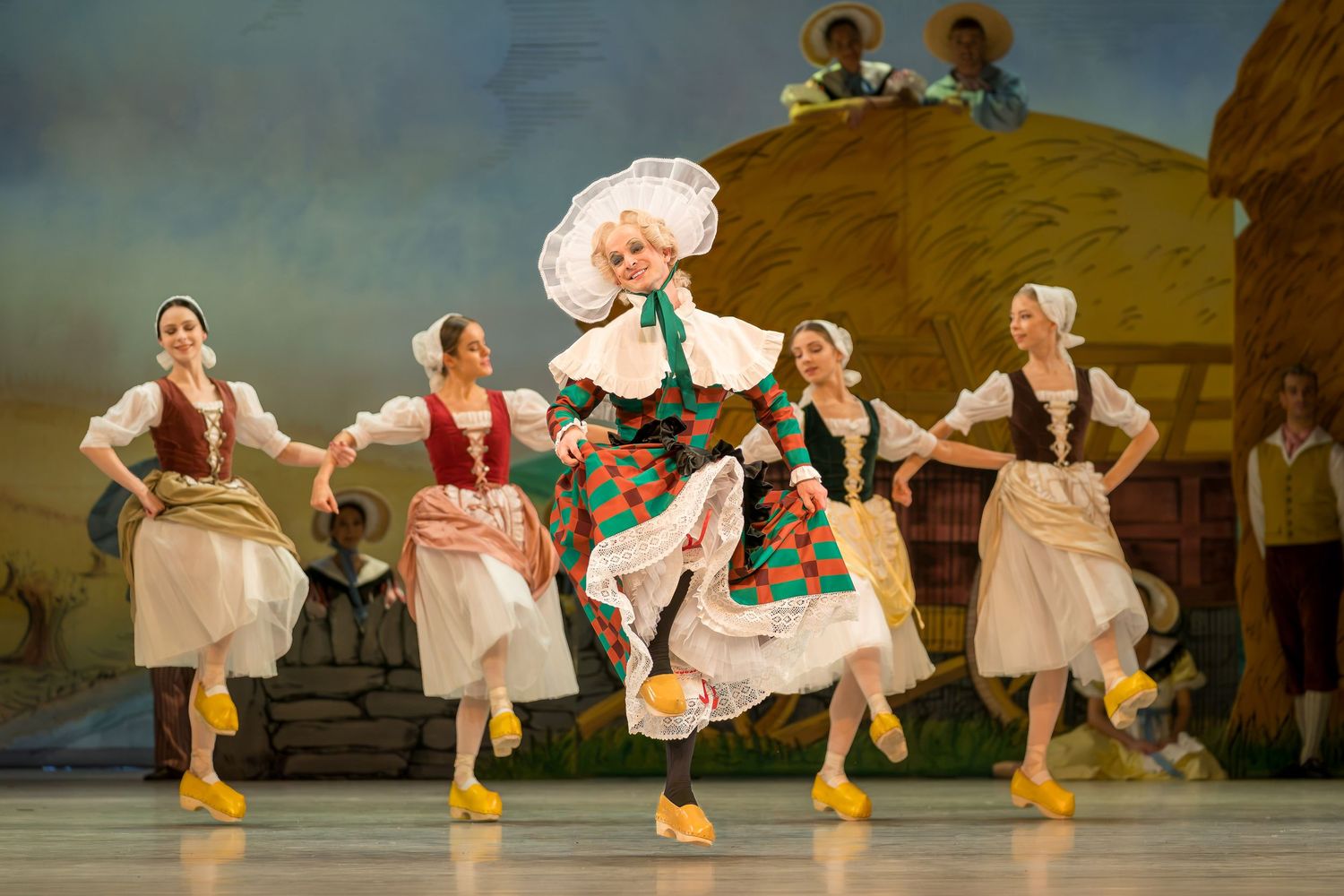 Five female dancers in traditional folk costumes perform a lively ballet dance on a colorful theatrical stage.