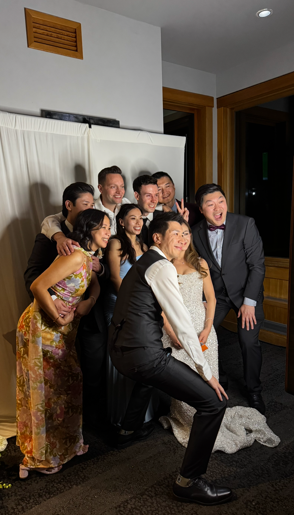 Group of friends posing joyfully at a wedding reception, with bride in white dress and guests in formal attire.