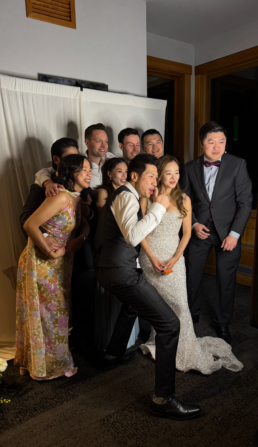 A joyful wedding party poses playfully in front of a white backdrop, with the bride in a sequined gown and groom in a tuxedo.