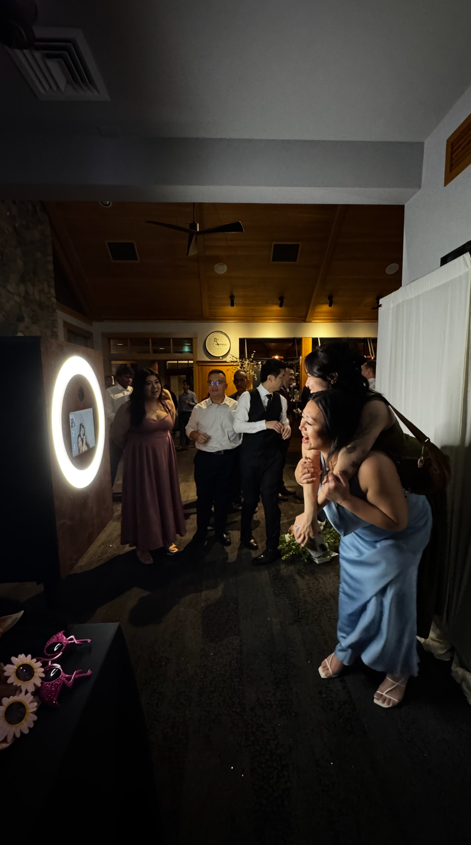 Guests in formal attire gather around a glowing ring light photo booth at an elegant indoor wedding reception.