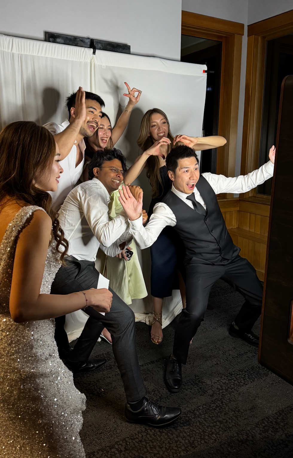 Group of wedding guests posing energetically and laughing together near a photo booth at a reception.