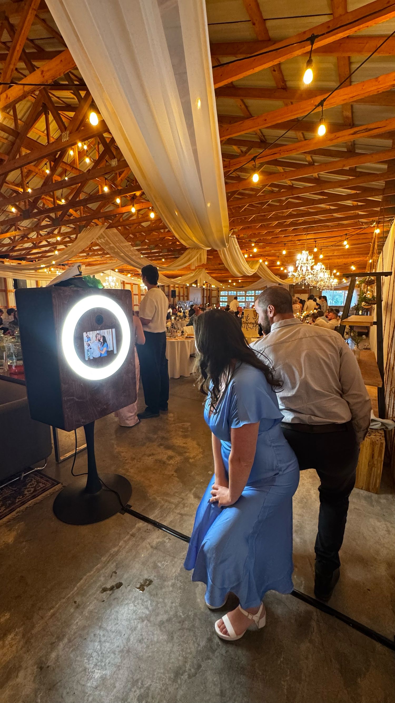Couple poses at a glowing ring light photo booth inside a rustic wedding venue with string lights and draped ceiling.