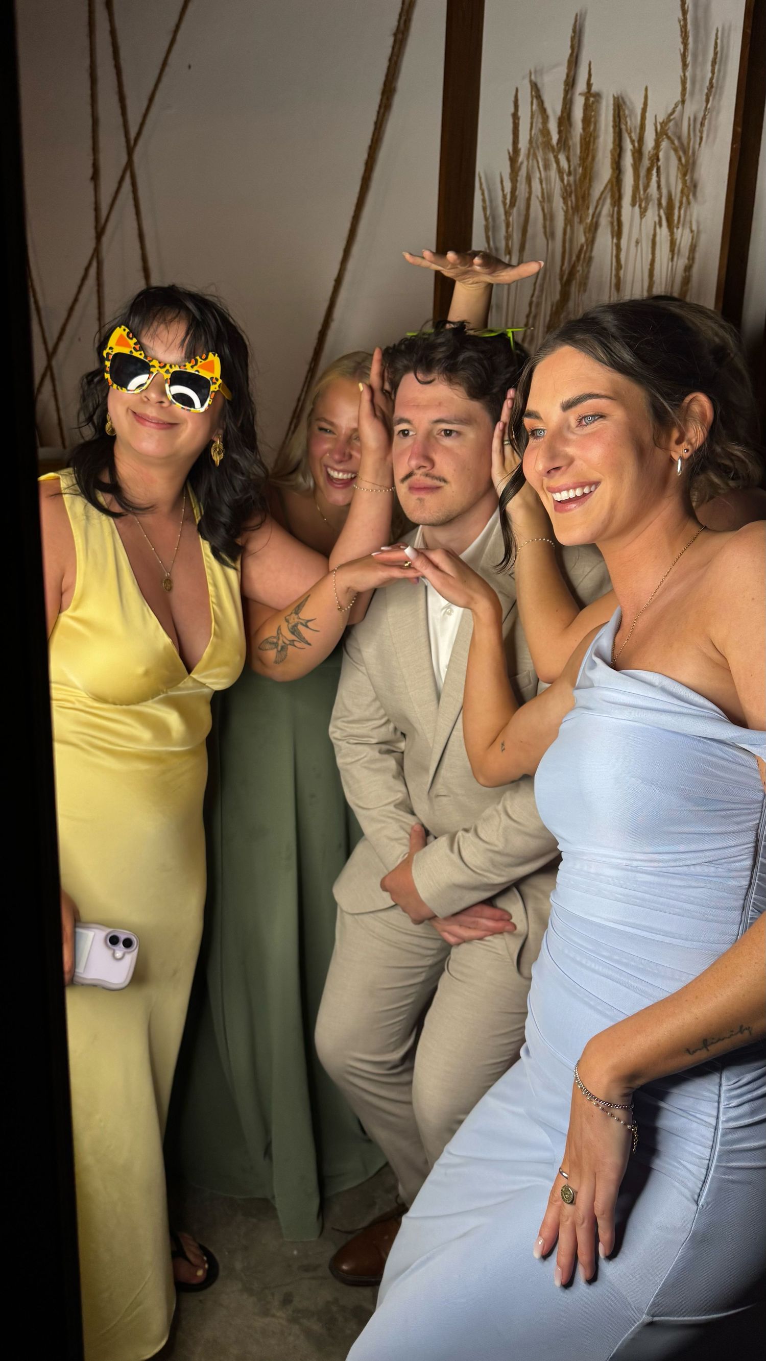 Four friends posing cheerfully at a wedding, wearing formal attire including yellow, sage, beige, and blue outfits.