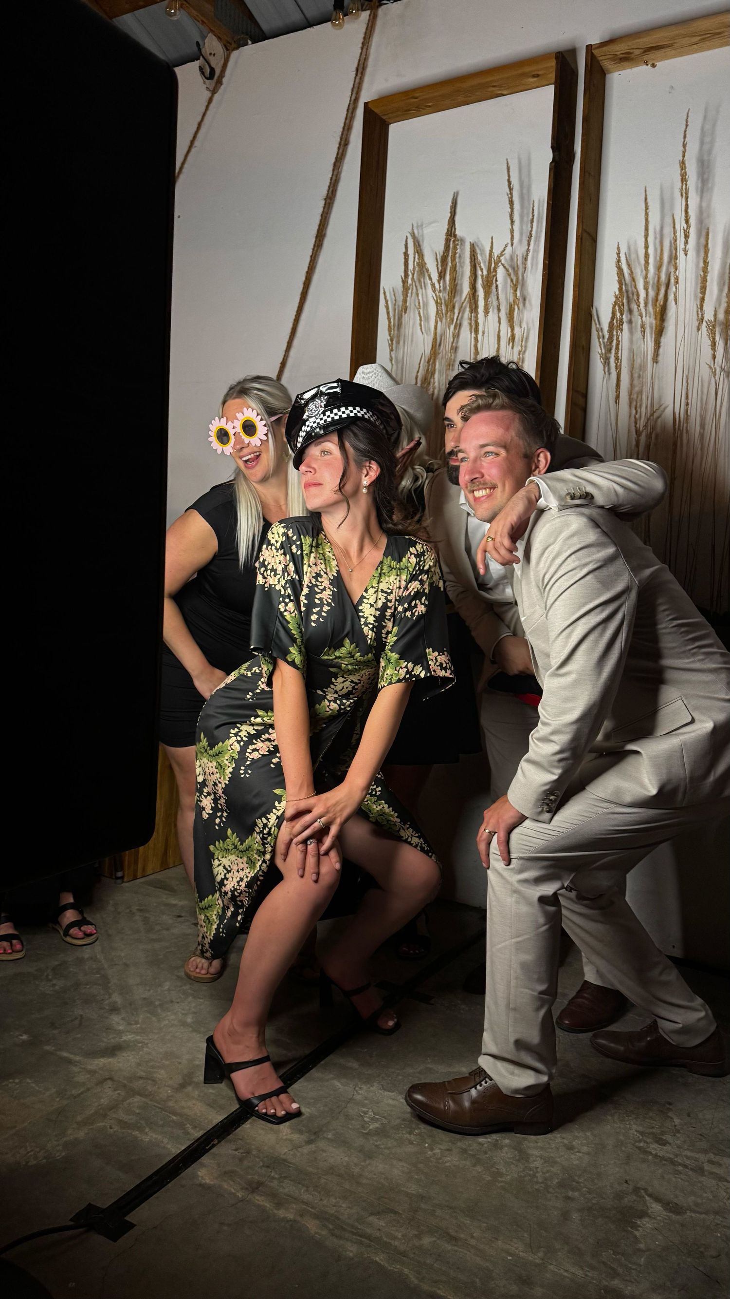 Three friends pose playfully at a party photo booth, wearing fun accessories including flower glasses and a police hat.