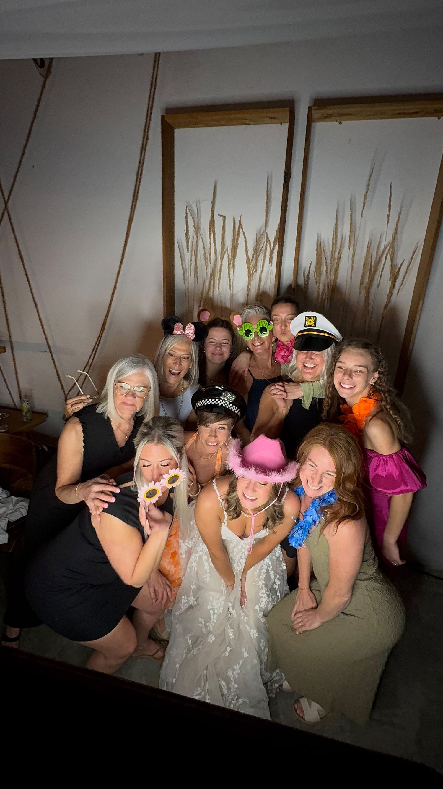A bride in a white gown poses with bridesmaids wearing fun photo booth props including hats and glasses at a wedding reception.