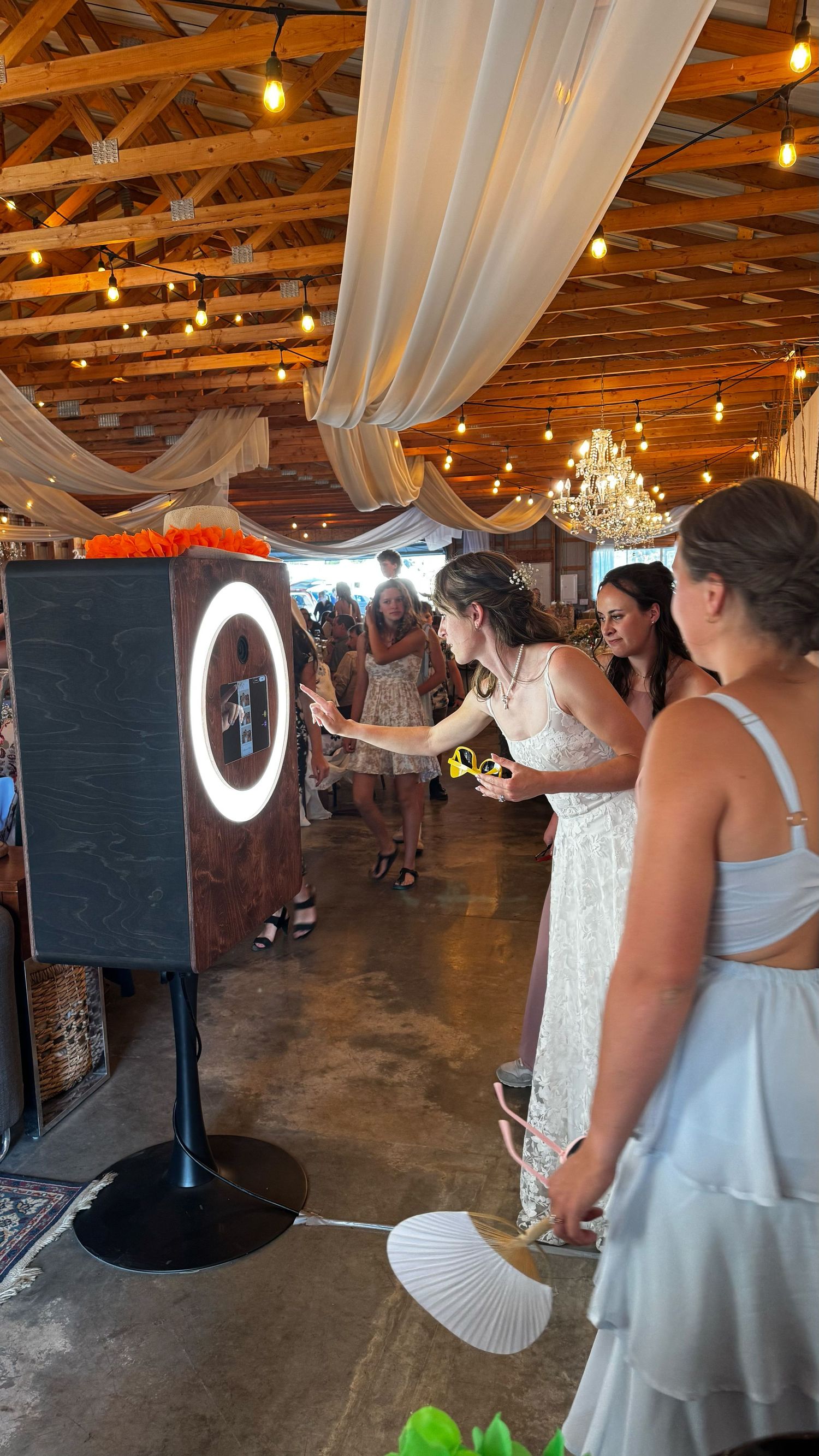 Bride and guests using a ring light photo booth at an elegant barn wedding reception with string lights and draped fabric.