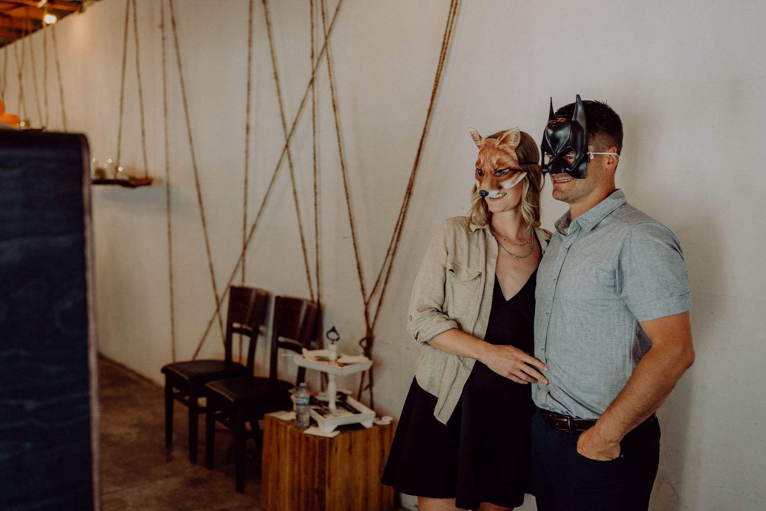 Couple wearing decorative masquerade masks posing together indoors at a rustic venue with rope swings and wooden chairs.