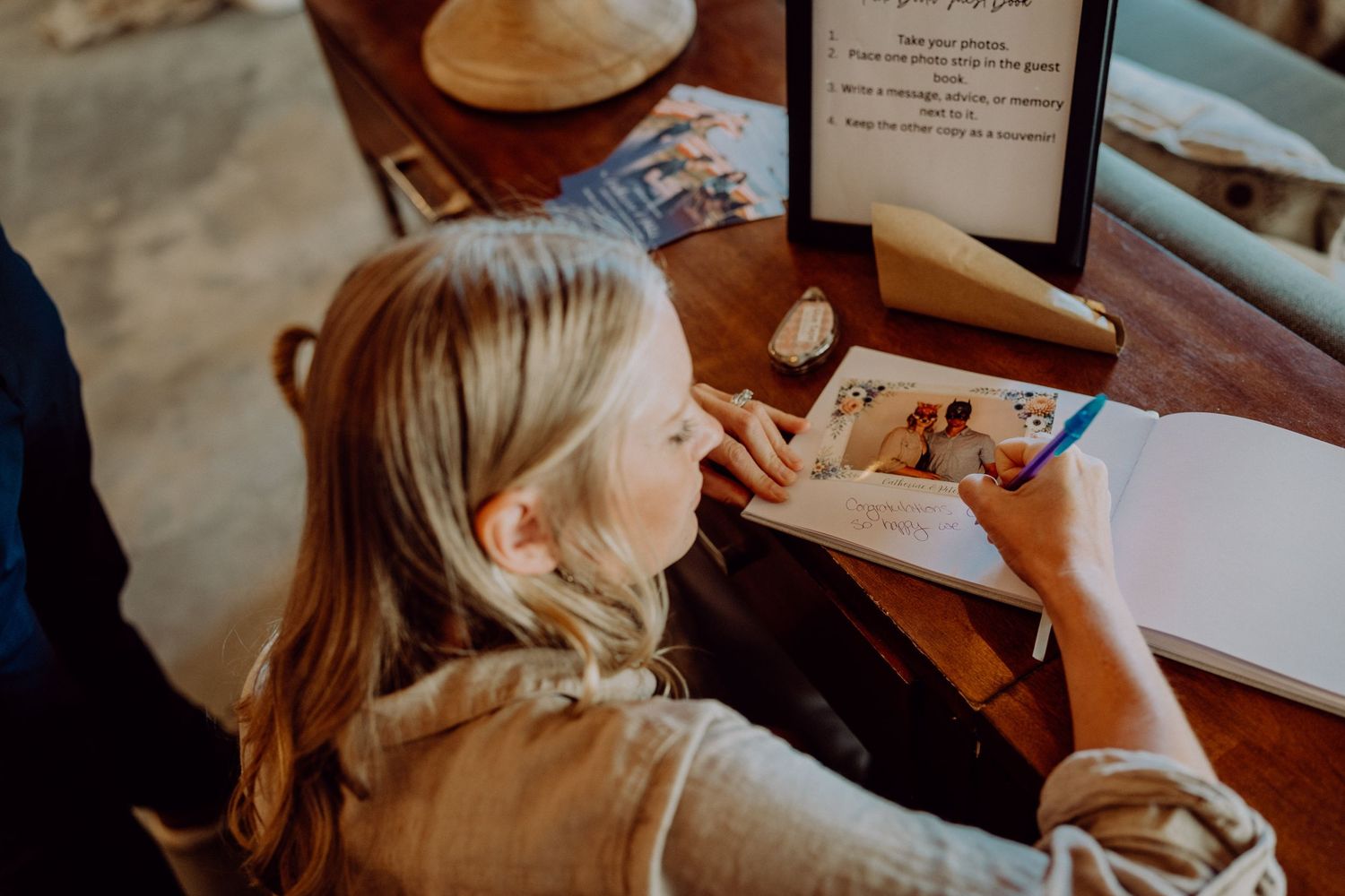 Blonde woman writing in a guest book with a blue pen at a wooden desk in a cozy interior setting.